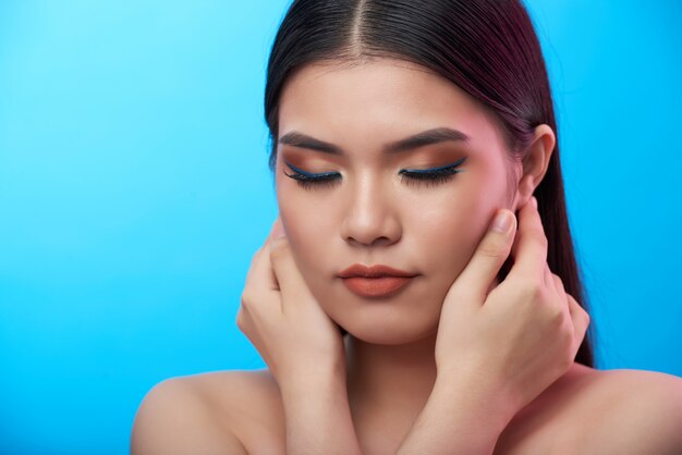 Closeup foto de joven asiática con maquillaje posando con los ojos cerrados y las manos tocando las mejillas