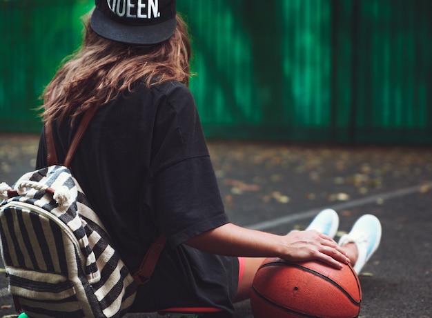Closeup foto pelota de baloncesto con niña sentada en plástico naranja penny shortboard sobre asfalto