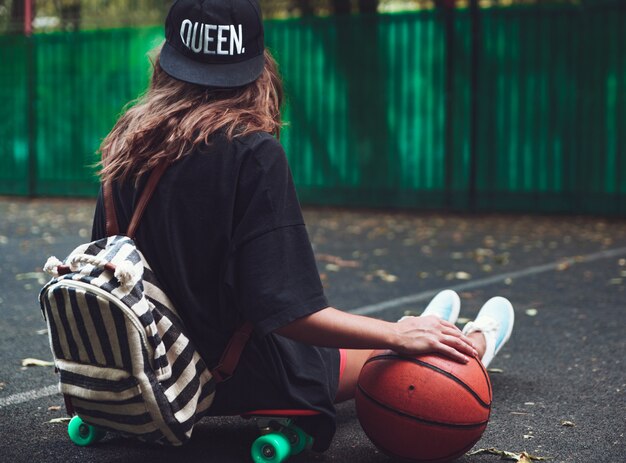 Closeup foto pelota de baloncesto con niña sentada en plástico naranja penny shortboard sobre asfalto