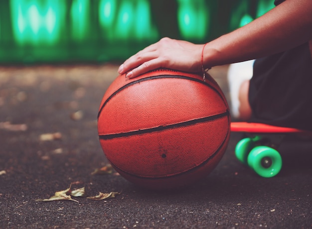 Closeup foto pelota de baloncesto con niña sentada en plástico naranja penny shortboard sobre asfalto