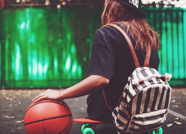 Closeup foto pelota de baloncesto con niña sentada en plástico naranja penny shortboard sobre asfalto