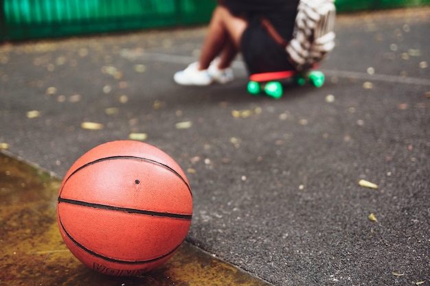 Closeup foto pelota de baloncesto con niña sentada en plástico naranja penny shortboard sobre asfalto