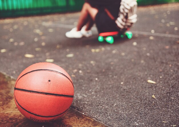 Closeup foto pelota de baloncesto con niña sentada en plástico naranja penny shortboard sobre asfalto