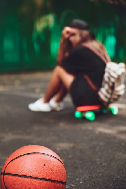 Closeup foto pelota de baloncesto con niña sentada en plástico naranja penny shortboard sobre asfalto