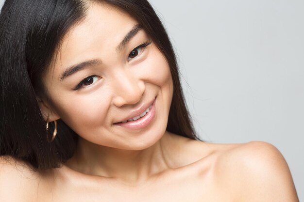 Closeup imagen de dama desnuda asiática alegre en estudio. Hermosa mujer con cabello negro mostrando sus hermosos hombros aislado sobre fondo gris.