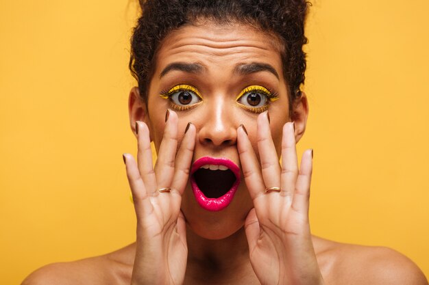 Closeup mujer afroamericana gritando emocionalmente o llamando a la cámara poniendo las manos en la boca aislada, sobre pared amarilla