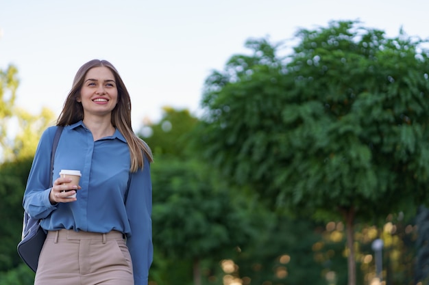 Closeup mujer atractiva en movimiento con café para llevar en las calles de la ciudad. Retrato chica rubia sosteniendo un vaso de papel con bebida caliente al aire libre.