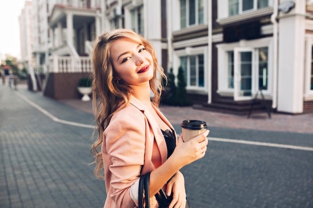 Closeup retrato atractivo modelo con labios vinosos caminando con café en chaqueta coral en la calle.