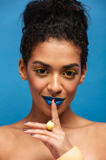 Closeup retrato de belleza de mujer africana semidesnuda con maquillaje de moda pidiendo guardar silencio o secreto poniendo el dedo en los labios aislados, sobre la pared azul