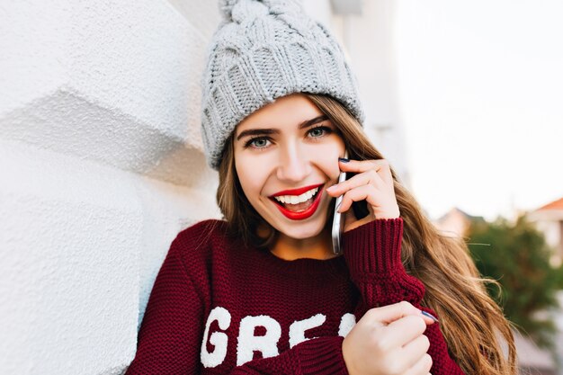Closeup retrato bonita morena con pelo largo con sombrero de punto y suéter marsala hablando por teléfono en la calle. Ella se ve emocionada.