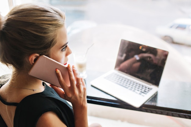 Foto gratuita closeup retrato de espalda hermosa mujer en vestido negro en cafetería. ella está hablando por teléfono, trabajando con una computadora portátil.