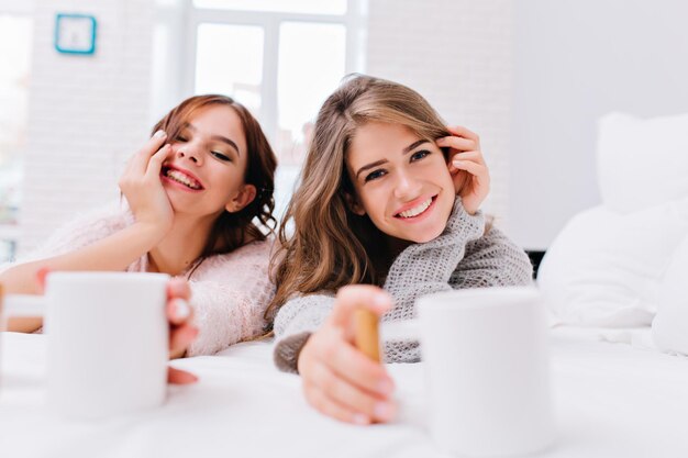 Closeup retrato feliz increíble chicas en suéteres de punto relajándose en la cama blanca con tazas de café. Mañana perfecta, emociones verdaderas, sonreír, divertirse.