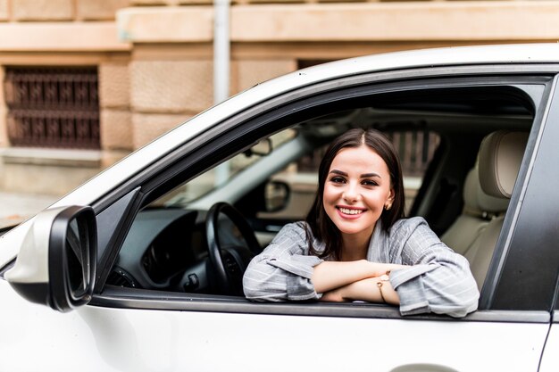 Closeup retrato feliz sonriente joven atractiva mujer comprador sentado en su nuevo coche emocionado listo para viaje aislado fuera de la oficina de lote de concesionario distribuidor. Concepto de compra de auto de transporte personal