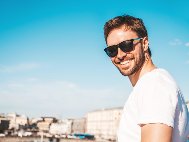 Foto gratuita closeup retrato de guapo sonriente hipster lambersexual modelo elegante hombre vestido con camiseta blanca moda hombre posando detrás de cielo azul en el fondo de la calle en gafas de sol