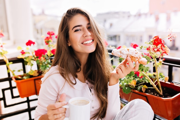 Closeup retrato hermosa chica con cabello largo desayunando en el balcón por la mañana en la ciudad. Ella sostiene una taza, croissant, sonriendo.