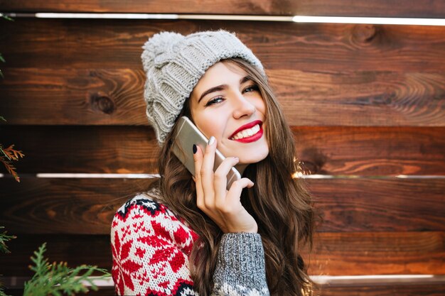 Closeup retrato hermosa chica con cabello largo y labios rojos en madera. Lleva un suéter y un gorro de invierno, habla por teléfono y sonríe.