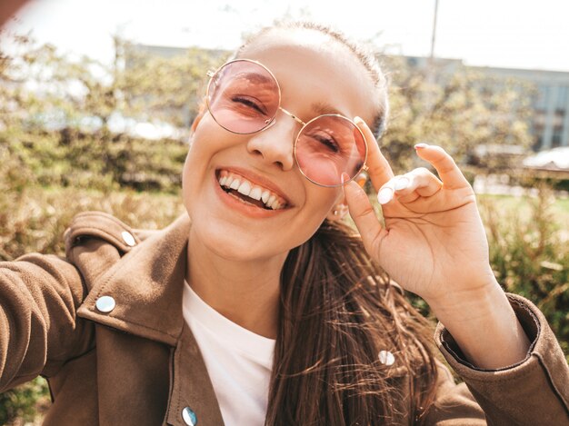 Closeup retrato de una hermosa niña morena sonriente en la chaqueta hipster de verano Modelo tomando selfie en teléfono inteligente Mujer haciendo fotos en un cálido día soleado en la calle con gafas de sol