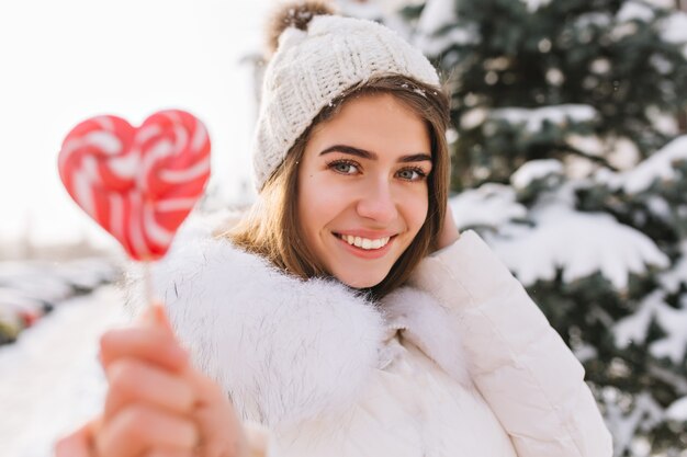 Closeup retrato increíble mujer sonriente alegre en la soleada mañana de invierno con piruleta rosa en la calle. Mujer joven atractiva con sombrero de lana blanco caliente disfrutando de un clima frío. Tiempo feliz, emociones positivas.