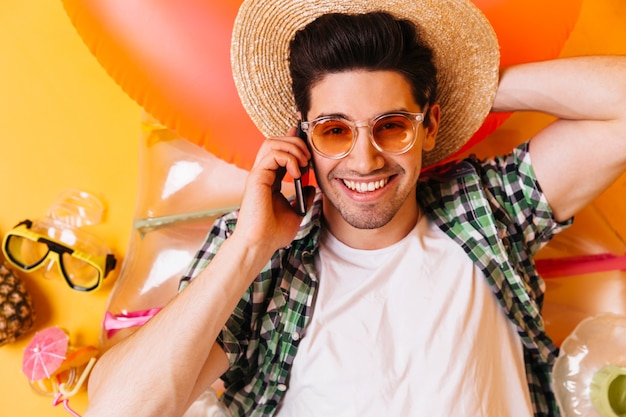 Foto gratuita closeup retrato de joven con gafas naranjas y sombrero de paja. el hombre en camiseta está descansando sobre un colchón inflable y hablando por teléfono.