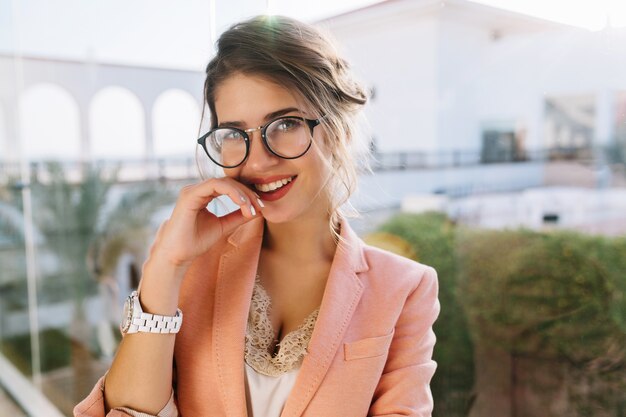 Closeup retrato de joven hermosa con gafas elegantes, bonita estudiante, mujer de negocios con elegante chaqueta rosa, blusa beige con encaje, maquillaje de día. Ventana grande con vista al patio.