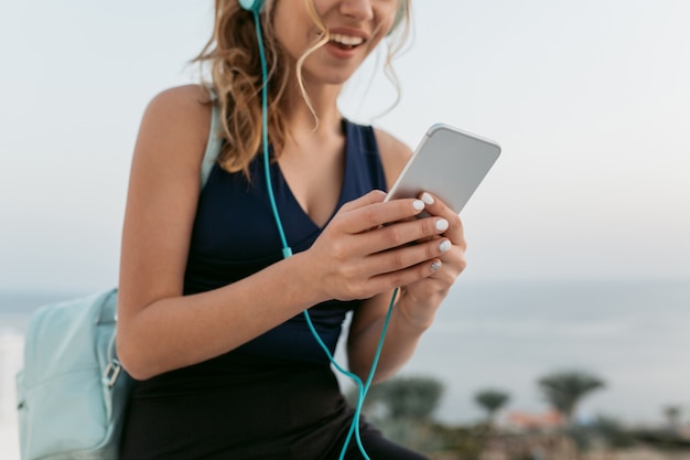 Closeup retrato de manos de mujer joven y bonita en ropa deportiva charlando por teléfono. disfrutar frente al mar, escuchar música a través de auriculares, divertirse, sonreír