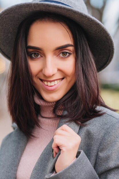 Closeup retrato de moda encantadora mujer joven con cabello morena, con sombrero gris sonriendo en la calle en la ciudad. Estado de ánimo alegre, verdaderas emociones positivas, hermosa modelo.