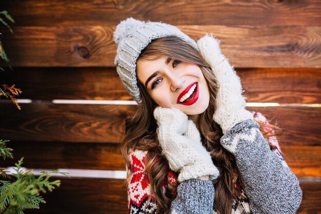 Closeup retrato morena hermosa chica con pelo largo en suéter de invierno y sombrero gris tejido en madera. Ella toca la cara con las manos en guantes y sonríe.