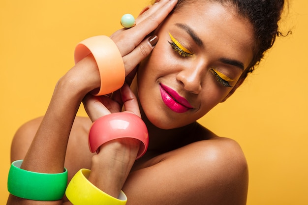 Closeup retrato de mujer afroamericana de moda con maquillaje brillante y accesorios multicolores tomados de la mano en la cara, aislado sobre la pared amarilla