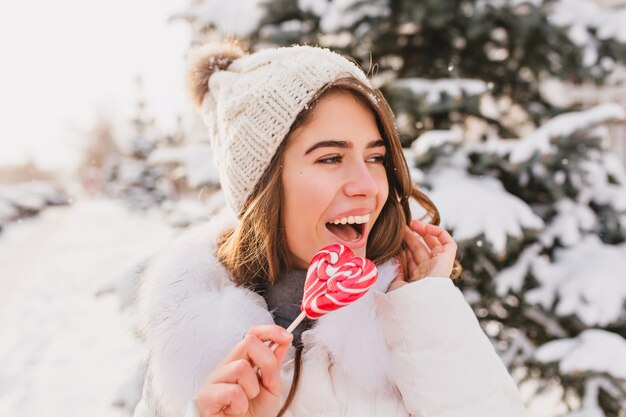 Closeup retrato mujer divertida linda con sombrero de lana blanco divirtiéndose con lollypop de corazón rosa en la calle. Bastante joven disfrutando del frío invierno, nieve, emociones brillantes.