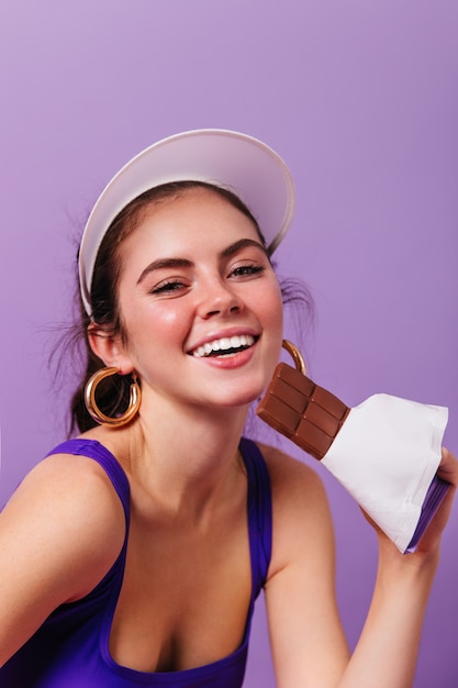 Closeup retrato de mujer joven en aretes de oro brillantes y gorra blanca sonriendo y sosteniendo la barra de chocolate en la pared púrpura