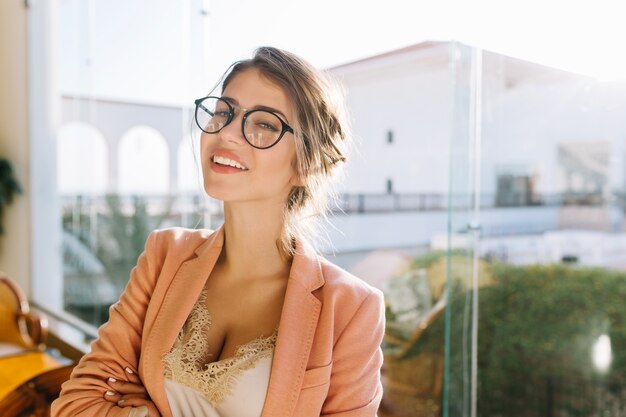 Closeup retrato de mujer joven con gafas elegantes, elegante dama en elegante chaqueta rosa con blusa beige, linda estudiante. Ventana grande con linda vista.