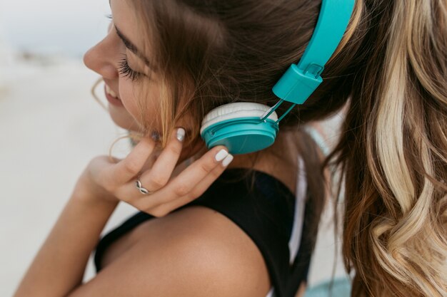 Closeup retrato de mujer joven con pelo largo y rizado disfrutando de una música encantadora a través de auriculares azules. Caminando por el paseo marítimo, sonriendo con los ojos cerrados, humor alegre