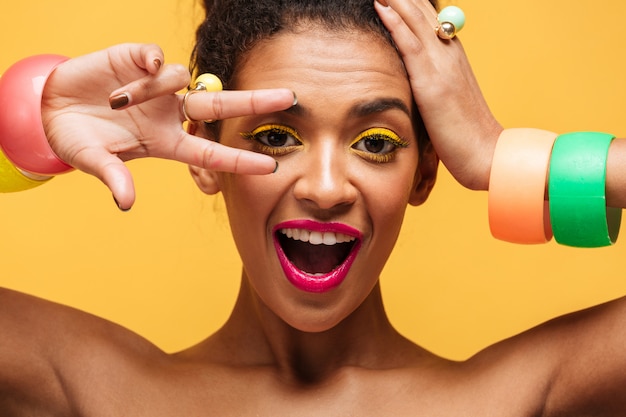 Closeup retrato de mujer mulata juguetona con párpados amarillos y labios rosados gesticulando dos dedos a los ojos y mirando a cámara, aislado