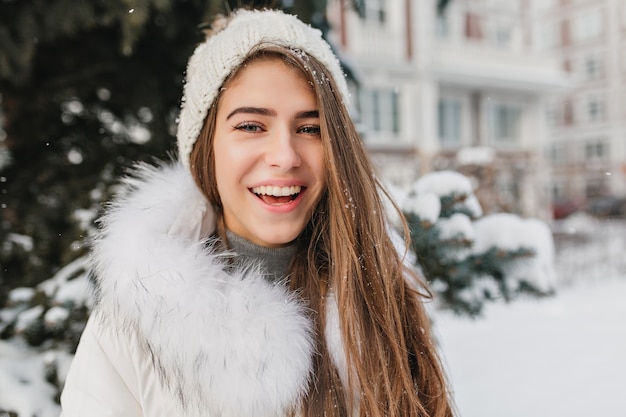 Closeup retrato de mujer sonriente alegre con gorro de punto posando al aire libre en la calle llena de nieve. Alegre dama rubia con ojos azules disfrutando del invierno pasando el fin de semana en el patio.