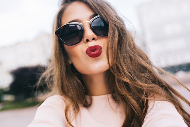 Closeup retrato de niña bonita en gafas de sol con peinado largo en la ciudad. Ella haciendo un beso con labios vinosos.