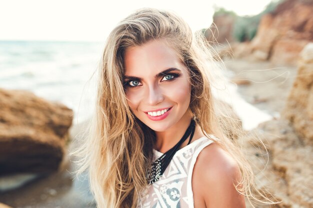 Closeup retrato de niña bonita rubia en la luz del sol posando en la playa rocosa. Ella esta sonriendo a la camara