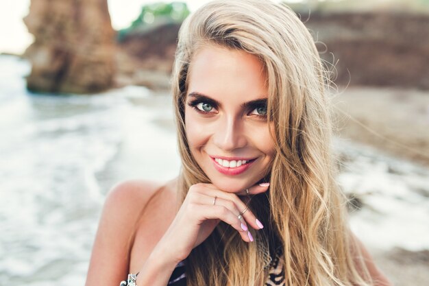 Closeup retrato de niña bonita rubia con pelo largo posando en la playa rocosa. Ella sonríe a la cámara.