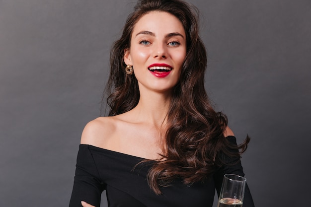 Closeup retrato de niña con ojos azules brillantes y lápiz labial rojo. Señora en top negro sonríe y sostiene una copa de vino blanco sobre fondo oscuro.