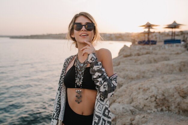 Closeup retrato de niña sonriente en la playa, puesta del sol. Vistiendo traje de baño negro de moda, bikini, elegantes gafas de sol, collar, chaqueta de punto, capa con adornos.