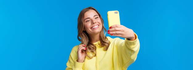 Closeup retrato tierna linda chica rubia en sudadera con capucha amarilla sosteniendo teléfono móvil tomando selfie con sma