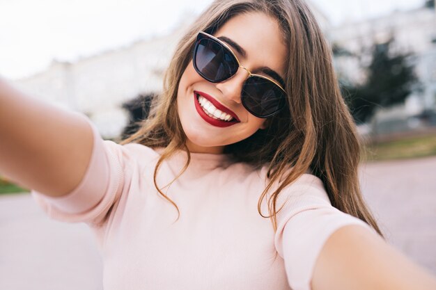 Closeup selfie-retrato de chica atractiva en gafas de sol con peinado largo y sonrisa blanca como la nieve en la ciudad.