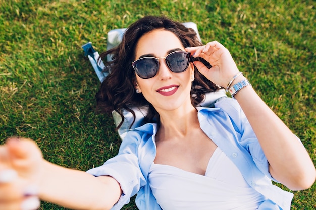 Closeup selfie-retrato de linda chica morena con pelo corto tendido sobre la hierba en el parque. Viste camiseta blanca y camisa azul, gafas de sol. Vista desde arriba.