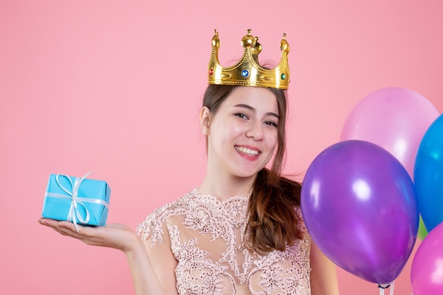 Closeup vista frontal feliz fiesta chica con corona sosteniendo presente y globos