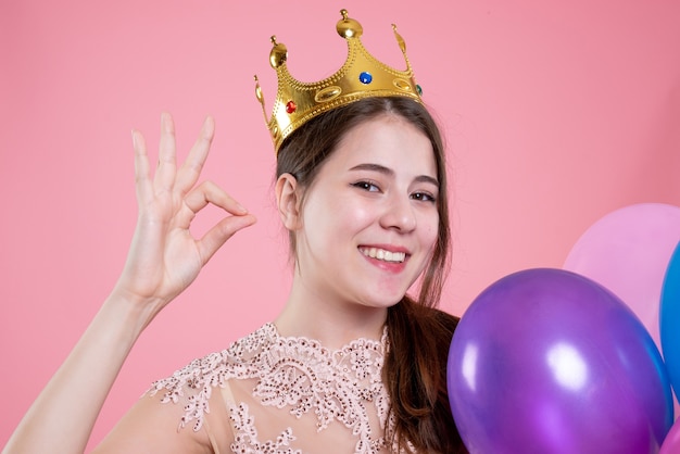 Closeup vista frontal linda chica fiestera con corona sosteniendo globos haciendo signo okey