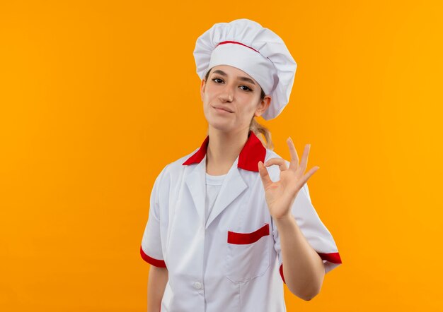 Cocinera bonita joven complacida en uniforme de chef haciendo signo ok aislado en el espacio naranja