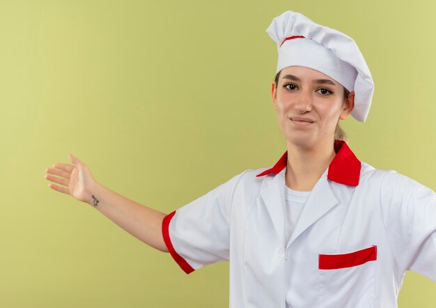 Cocinera bonita joven complacida en uniforme de chef mirando con el brazo abierto aislado en el espacio verde