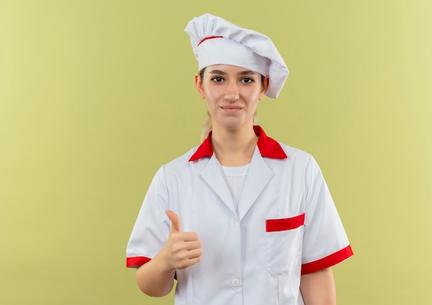 Cocinera bonita joven complacida en uniforme de chef mostrando el pulgar hacia arriba aislado en el espacio verde