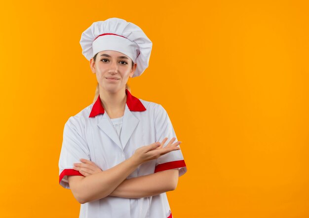 Cocinera bonita joven complacida en uniforme de chef de pie con postura cerrada y mostrando la mano vacía aislada en el espacio naranja