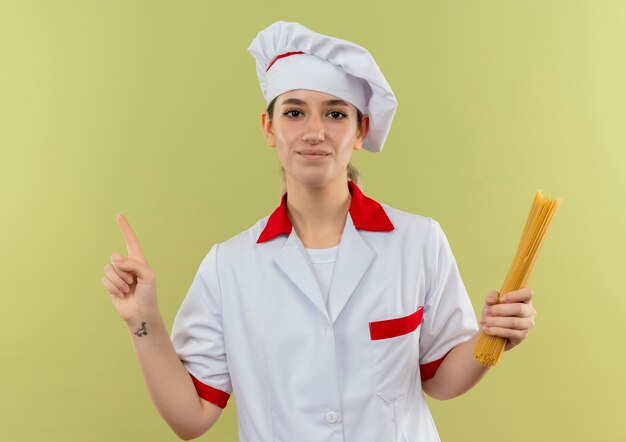 Cocinera bonita joven complacida en uniforme de chef sosteniendo pasta de espagueti y levantando el dedo aislado en espacio verde