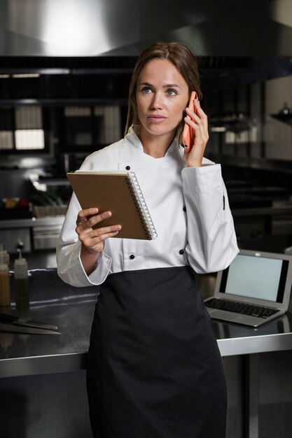 Cocinera en la cocina hablando por teléfono y usando un portátil